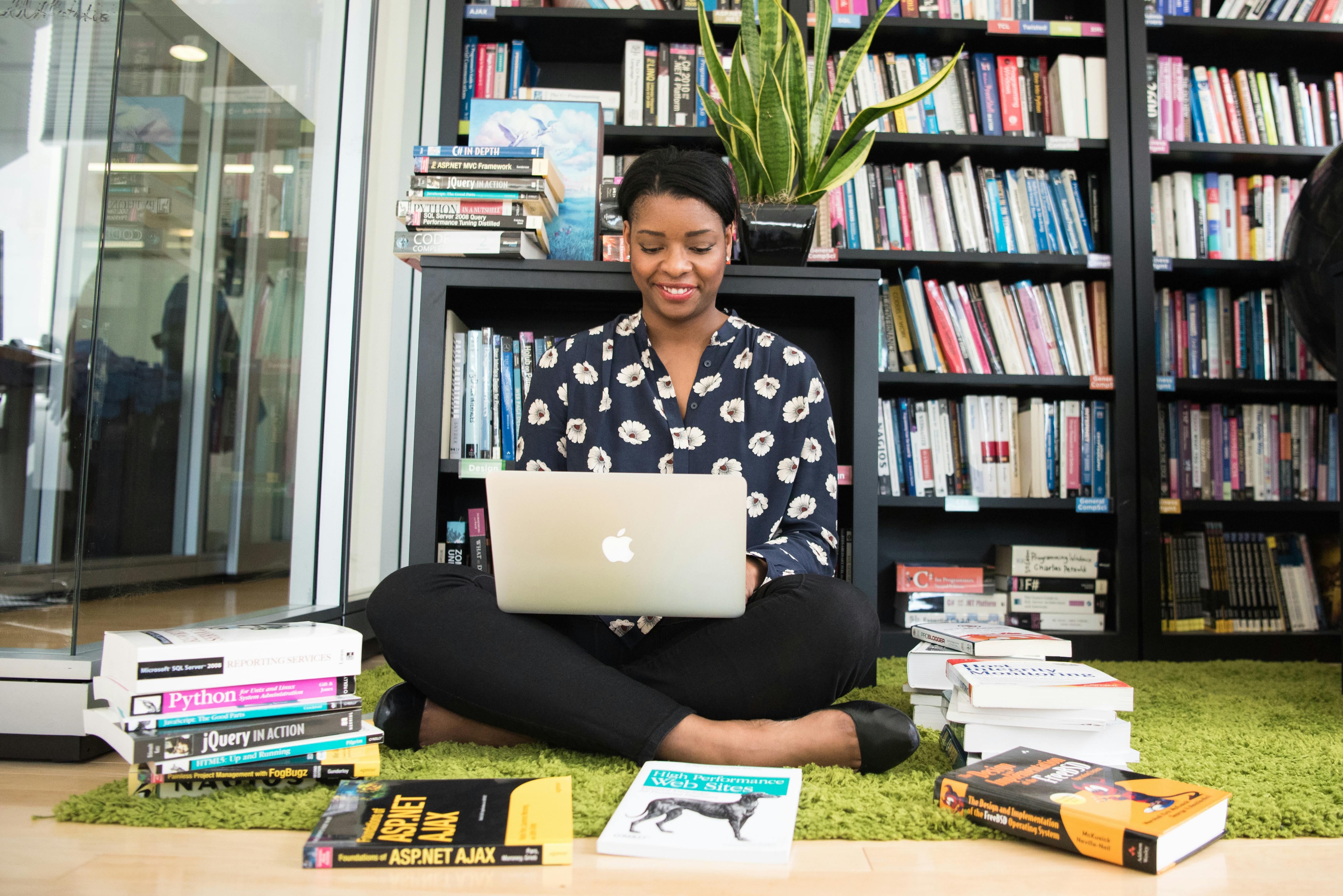 a girl sitting in an office on the floor surrounded by books and typing on a laptop